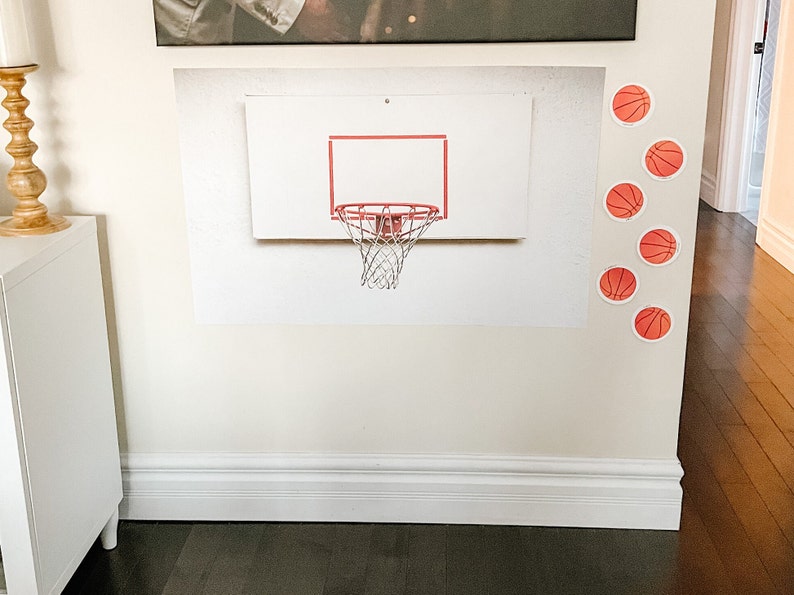May include: A white basketball hoop with a red rim and a black net is mounted on a white wall. There are several orange basketballs with black outlines on the wall around the hoop.