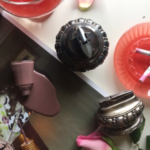 May include: Two silver cigarette lighters, one with a round top and the other with a more rectangular shape. Both lighters have intricate designs and are in a vintage style.