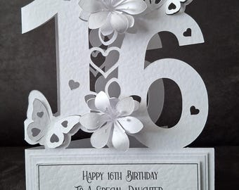 May include: White paper card with a 3D design of the number 16. The number is decorated with white paper flowers and butterflies. The card has a message that reads "Happy 16th Birthday To A Special Daughter Maria".