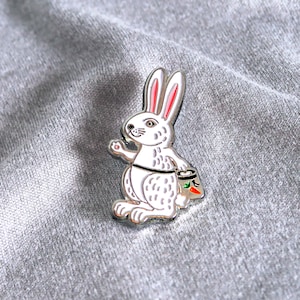 Puede incluir: Un pin de esmalte blanco de un conejo que lleva una zanahoria en una bolsa pequeña. El conejo tiene orejas rosas y un cuerpo blanco con ojos negros.