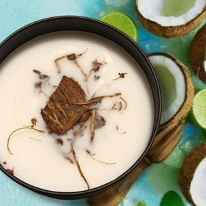 May include: A black bowl filled with a creamy white liquid topped with a central piece of brown bark and thin woody fibers. The bowl is surrounded by halved brown coconuts with white flesh, bright green lime slices, and green mint leaves.