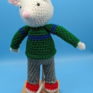 May include: A white crocheted mouse wearing a green and blue striped sweater and gray pants with red shoes. The mouse has a pink nose and black eyes.