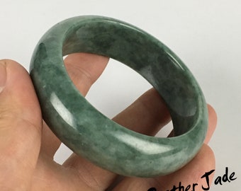 May include: A green jade bangle bracelet. The polished surface of the bracelet reflects light, highlighting the natural variations in the stone. The bracelet is a solid, circular design, suitable for everyday wear.
