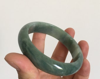 May include: A green jade bangle bracelet with a smooth, polished finish. The bangle is a light green colour with darker green swirls and streaks throughout.