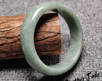 May include: A light green jade bangle bracelet. The bangle is smooth and polished, with a subtle swirl pattern. The bangle is resting on a piece of wood.