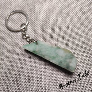 Puede incluir: Un llavero de piedra de jade verde con un anillo de llave de plata. La piedra es de color verde claro con vetas blancas que la atraviesan. El llavero está unido a una cadena de plata.