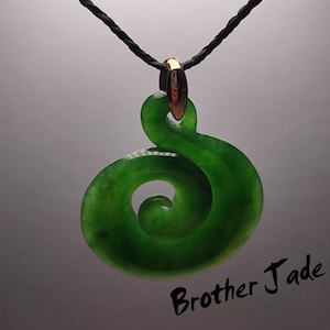 May include: A green jade pendant with a spiral design, hanging from a black cord with a gold clasp. The pendant is a traditional Maori symbol called a Koru, which represents new beginnings and growth.