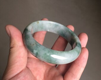 May include: A green jade bangle bracelet with a polished finish. The bracelet is a light green colour with darker green swirls throughout.