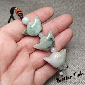 Puede incluir: Un colgante de jade con tres dijes de cabeza de zorro estilizadas en tonos verdes y blancos, unidos por pequeñas cuentas blancas. El colgante está suspendido de un cordón marrón y negro. El texto "Brother Jade" es visible.