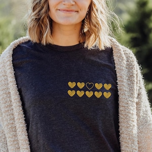 A smiling woman stands outdoors wearing a dark t-shirt adorned with ten yellow heart graphics, paired with a cozy beige cardigan. Green trees are visible in the background, suggesting a natural setting.