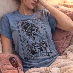 May include: A light blue t-shirt with a black graphic of a skeleton drinking from a cup, with the words "FLARE DAY". The person is holding a pink heating pad with a remote.