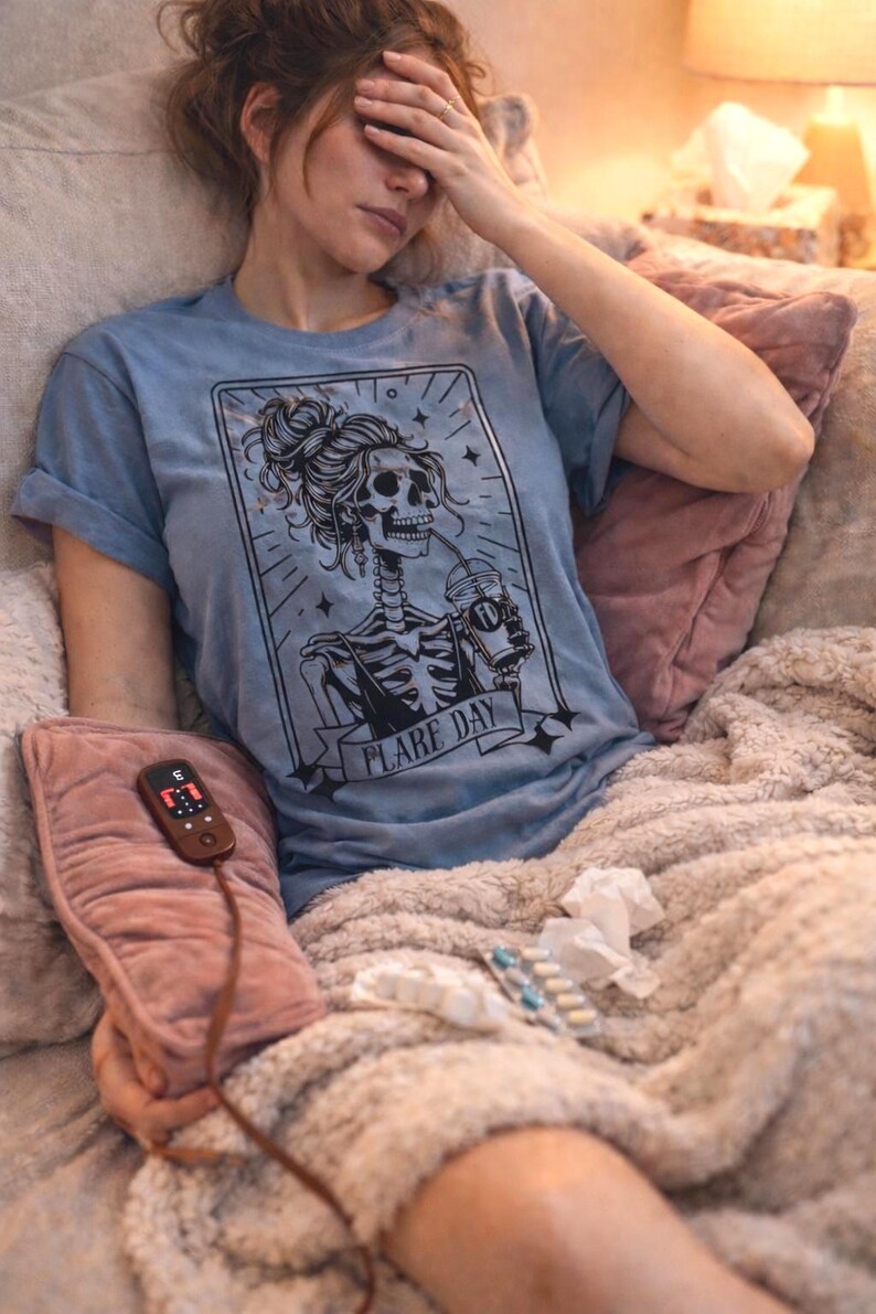 May include: A light blue t-shirt with a black graphic of a skeleton drinking from a cup, with the words "FLARE DAY". The person is holding a pink heating pad with a remote.