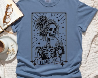 Puede incluir: Camiseta azul vaquero con un gráfico negro de una mujer esqueleto bebiendo de una taza. El esqueleto tiene un peinado de moño y está enmarcado por un borde decorativo. El texto "FLARE DAY" está debajo de la imagen.