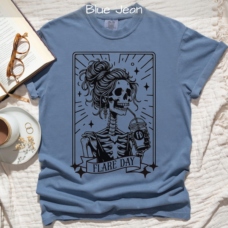 A blue jean colored t-shirt featuring a skeleton woman with a stylish hair bun, holding a drink, and the text 'FLARE DAY' in a tarot card design. The scene is laid on a textured background with sunglasses, a book, and a cup.