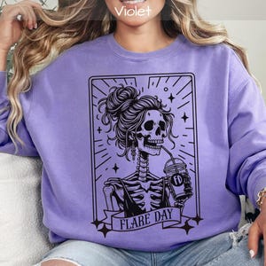May include: A violet sweatshirt featuring a black graphic of a skeleton with a bun, drinking from a cup. The skeleton is inside a rectangular frame with the words "FLARE DAY" on a banner below. The sweatshirt is a solid violet color.
