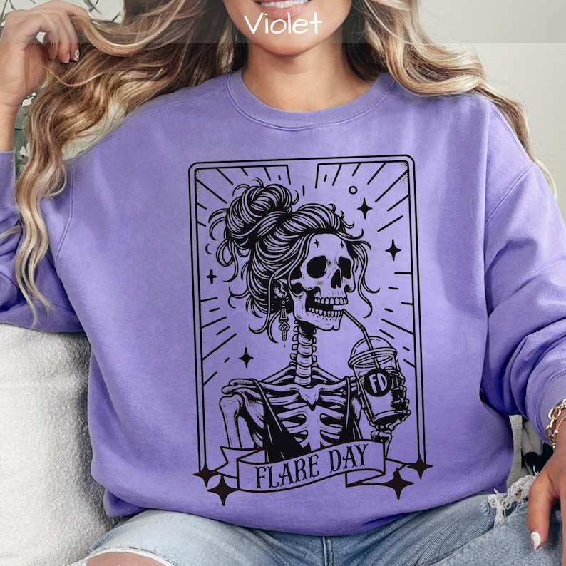 May include: A violet sweatshirt featuring a black graphic of a skeleton with a bun, drinking from a cup. The skeleton is inside a rectangular frame with the words "FLARE DAY" on a banner below. The sweatshirt is a solid violet color.