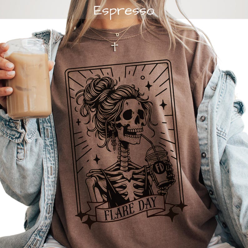 May include: Espresso-colored t-shirt featuring a black graphic of a skeleton with a bun, drinking from a cup. The design is framed with lines and stars, and the text "FLARE DAY" is on a banner below. The shirt is worn with a denim jacket.