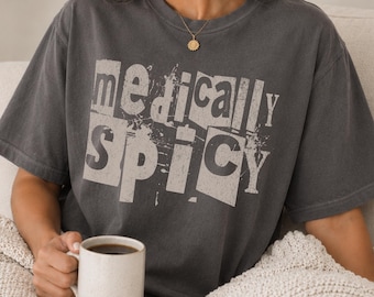 May include: A charcoal gray short-sleeve t-shirt featuring the text "medically spicy" in a distressed, block-letter style. The lettering appears in a light, off-white color with a weathered, textured finish across the chest of the garment.