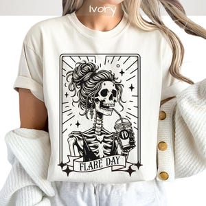 May include: Ivory-colored t-shirt featuring a black and white graphic of a skeleton with a messy bun, drinking from a cup. The graphic is framed with stars and the words "FLARE DAY" on a banner. The shirt has short sleeves.