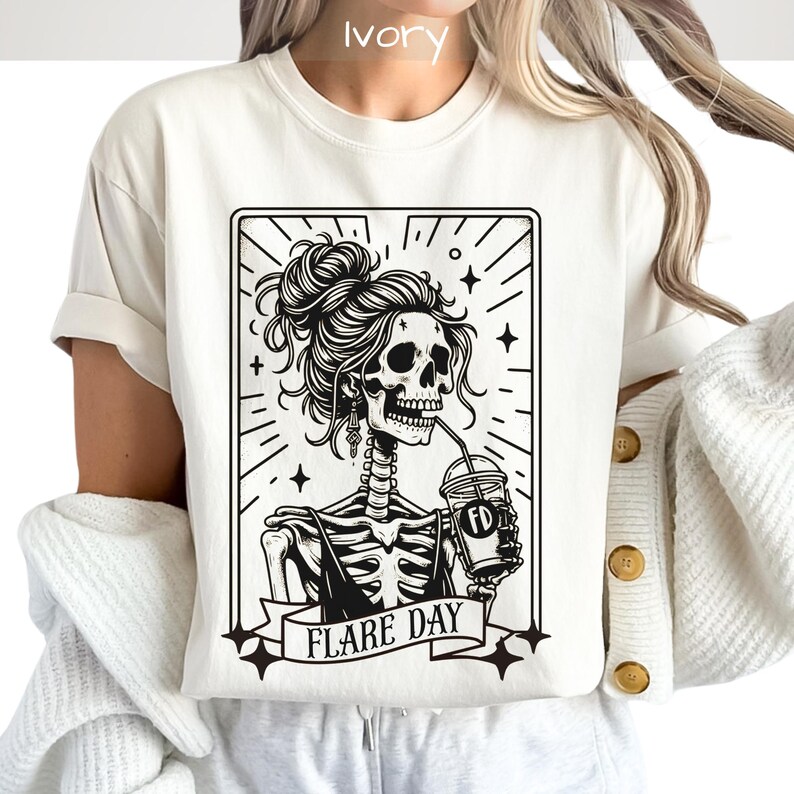 May include: Ivory-colored t-shirt featuring a black and white graphic of a skeleton with a messy bun, drinking from a cup. The graphic is framed with stars and the words "FLARE DAY" on a banner. The shirt has short sleeves.