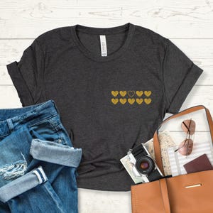 May include: A dark heather grey t-shirt with a row of gold hearts, one of which is black. The shirt is made of a soft, comfortable material and has a relaxed fit.