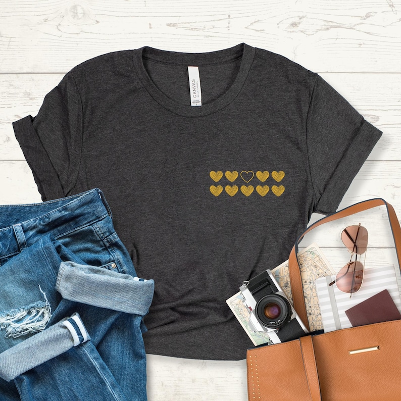 May include: A dark heather grey t-shirt with a row of gold hearts, one of which is black. The shirt is made of a soft, comfortable material and has a relaxed fit.