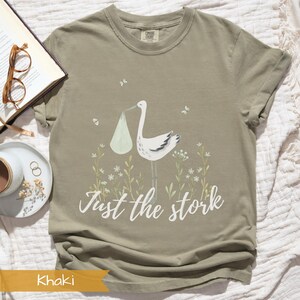 May include: Khaki t-shirt with a graphic of a stork carrying a baby in a white bag. The text "Just the stork" is printed below the graphic.
