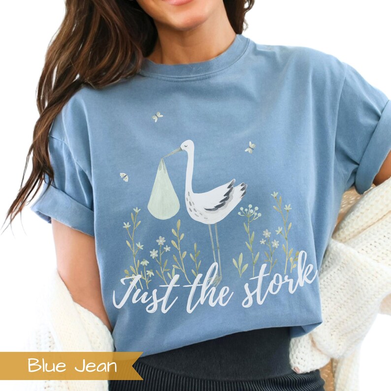 May include: A blue jean t-shirt with a white stork carrying a baby in a white bag. The text "Just the stork" is printed in white below the stork.