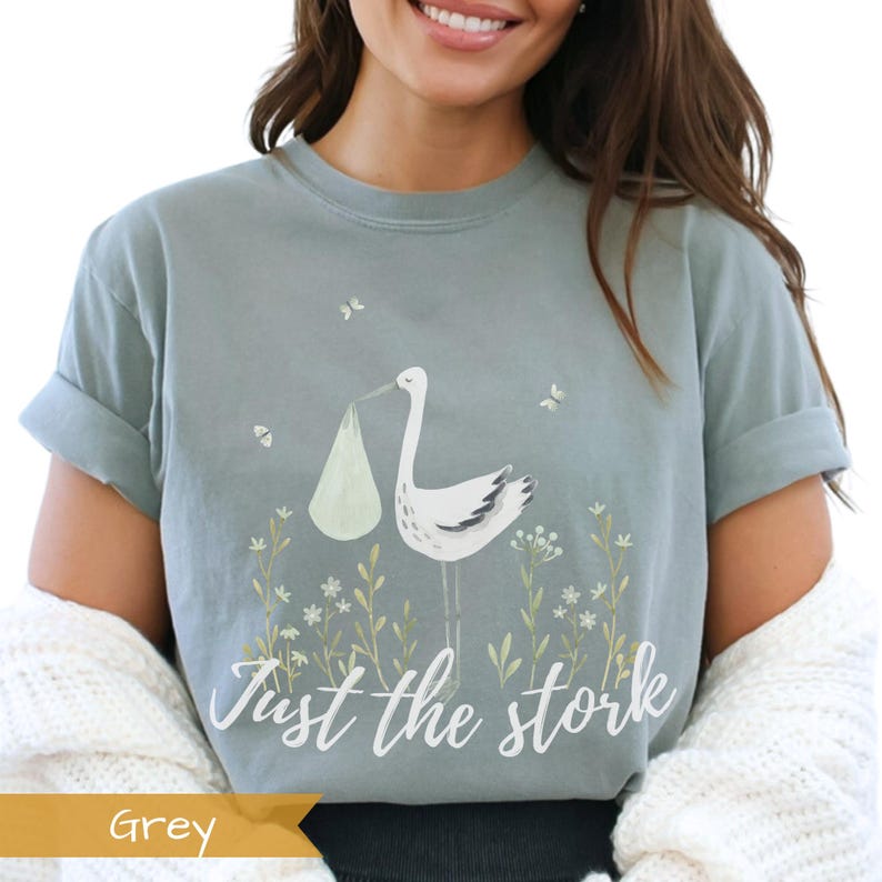 May include: A light blue t-shirt with a white stork carrying a baby in a green bundle. The shirt has the text "Just the stork" printed on it.