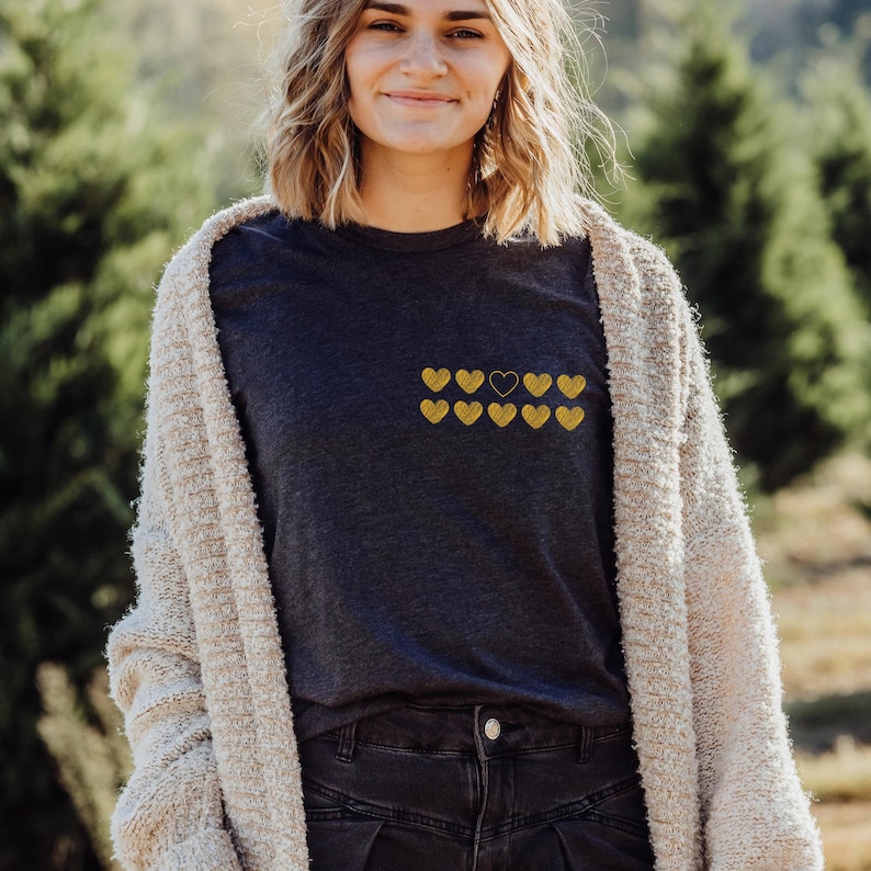 A smiling woman stands outdoors wearing a dark t-shirt adorned with ten yellow heart graphics, paired with a cozy beige cardigan. Green trees are visible in the background, suggesting a natural setting.