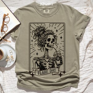 May include: Khaki-colored t-shirt featuring a black and white graphic of a skeleton with a bun, drinking from a cup. The graphic includes the text "FLARE DAY". The shirt is made of a soft material.