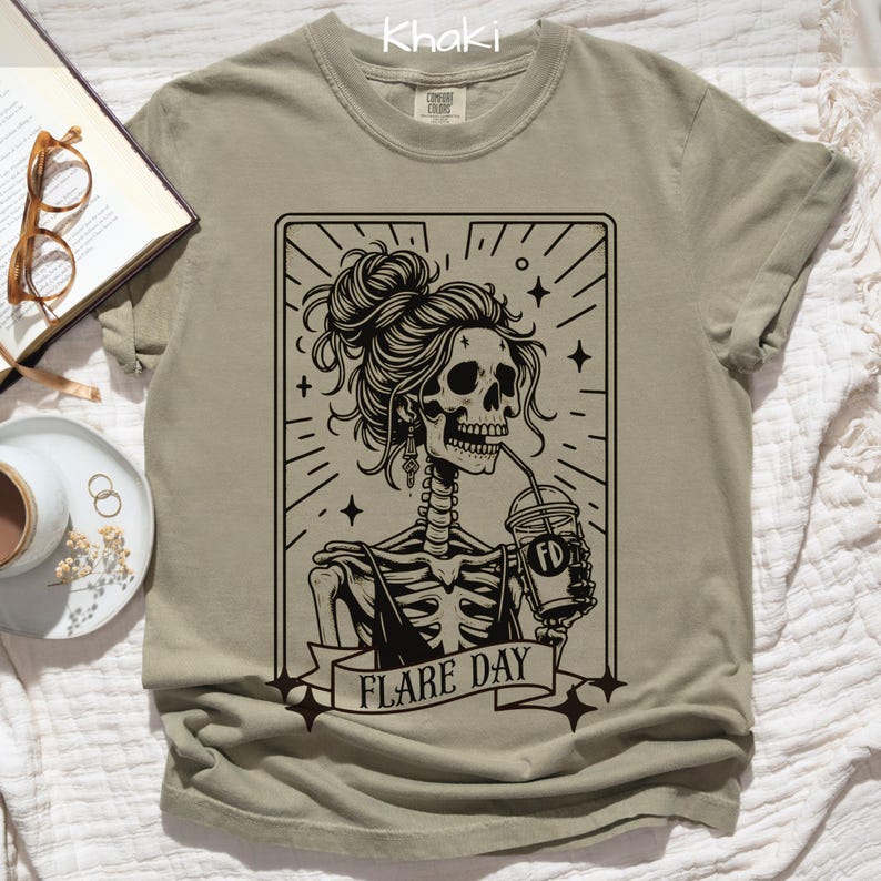 May include: Khaki-colored t-shirt featuring a black and white graphic of a skeleton with a bun, drinking from a cup. The graphic includes the text "FLARE DAY". The shirt is made of a soft material.