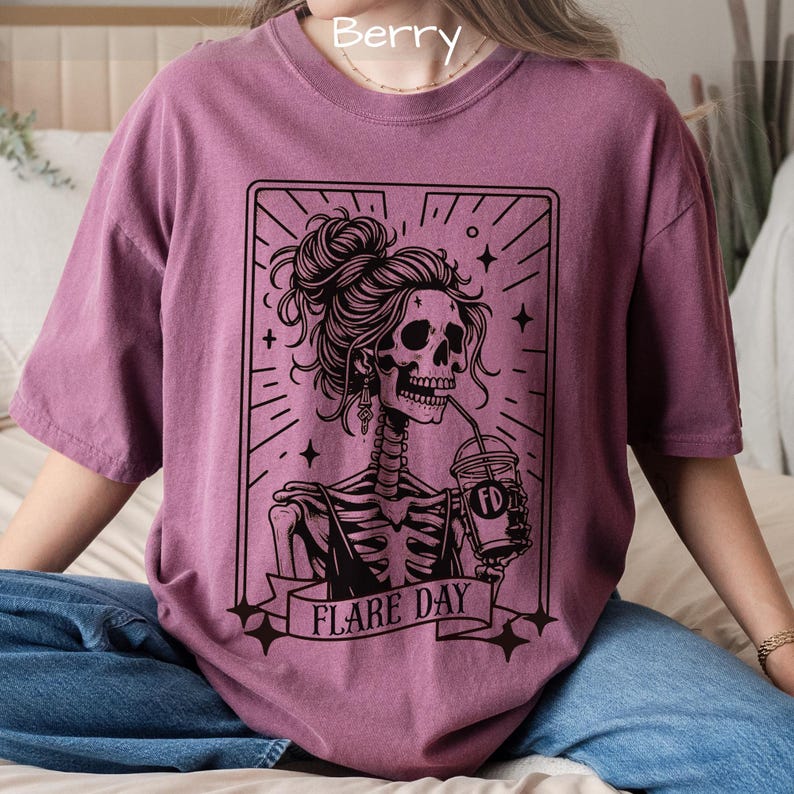 May include: Berry-colored t-shirt featuring a black graphic of a skeleton woman with a bun, drinking from a cup. The graphic is framed with lines and stars, and the text "FLARE DAY" is displayed on a banner below the skeleton.