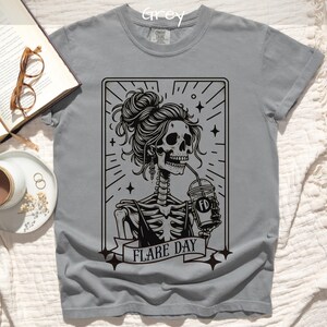 May include: Grey t-shirt featuring a black graphic of a skeleton woman with a messy bun, drinking from a coffee cup. The text "FLARE DAY" is on a banner below the image. The shirt is a crew neck.