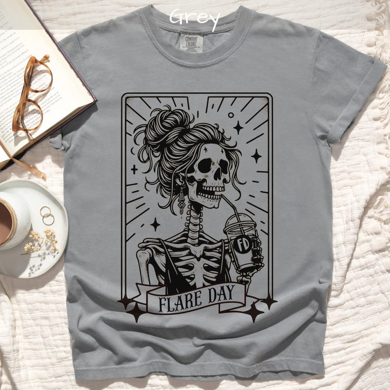 May include: Grey t-shirt featuring a black graphic of a skeleton woman with a messy bun, drinking from a coffee cup. The text "FLARE DAY" is on a banner below the image. The shirt is a crew neck.