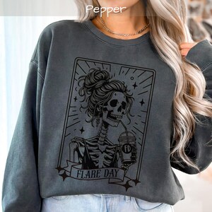 May include: Dark gray sweatshirt with a black graphic of a skeleton with a messy bun, drinking from a cup. The graphic is framed with stars and lines, and the words "FLARE DAY" are on a banner below. The word "Pepper" is at the top.