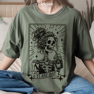 May include: Moss green t-shirt featuring a black graphic of a skeleton with a bun hairstyle, drinking from a cup. The graphic is framed with radiating lines and stars, with the words "FLARE DAY" on a banner.