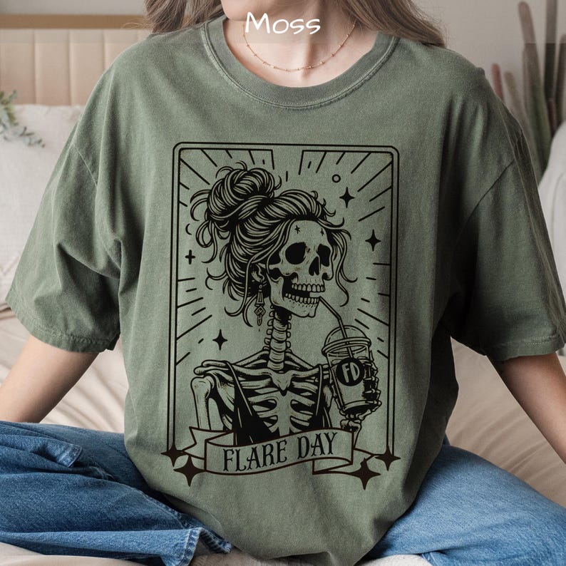 May include: Moss green t-shirt featuring a black graphic of a skeleton with a bun hairstyle, drinking from a cup. The graphic is framed with radiating lines and stars, with the words "FLARE DAY" on a banner.