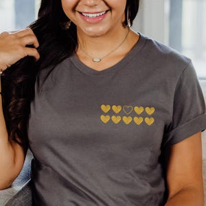May include: A gray t-shirt with a gold embroidered design of nine hearts, one of which is larger than the others.