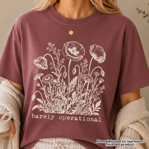 May include: A maroon short-sleeved t-shirt featuring a white line-art illustration of various wildflowers and poppies. The text "barely operational" is printed in a white, lowercase, handwritten-style font centered below the floral graphic.