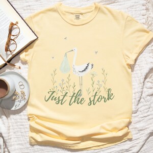 May include: A yellow t-shirt with a white stork carrying a baby in a green bag. The text "Just the stork" is printed in green below the stork.