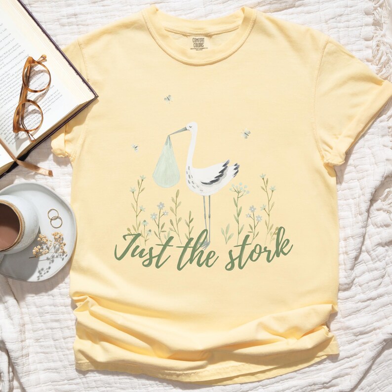 May include: A yellow t-shirt with a white stork carrying a baby in a green bag. The text "Just the stork" is printed in green below the stork.