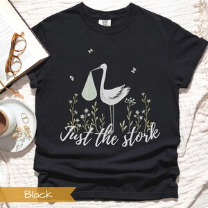 May include: Black t-shirt with a white stork carrying a baby in a white sack. The stork is surrounded by green leaves and white flowers. The text "Just the stork" is printed below the stork.