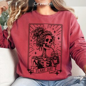 May include: Crimson-colored sweatshirt with a black graphic of a skeleton with a messy bun, drinking from a cup. The graphic includes the text "FLARE DAY" on a banner. The sweatshirt has a crew neck and long sleeves.