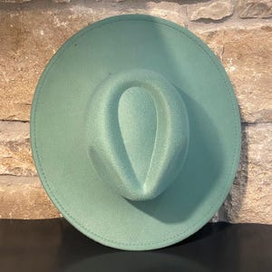 May include: A wide-brimmed hat in a muted green color. The hat has a slightly indented crown and a stitched brim.