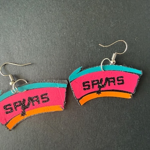 May include: A pair of handmade earrings featuring a pink, teal, and orange design with the word "SPURS" in black glitter lettering. The earrings have silver-colored fish hook ear wires.