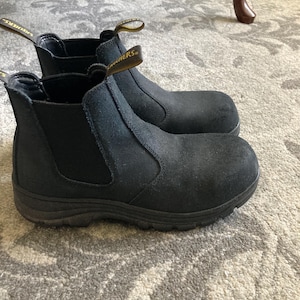 May include: A pair of black suede Chelsea boots with elastic side panels and a thick rubber sole. Each boot features a yellow and black pull tab at the heel with the Skechers brand name printed in black text on the fabric.