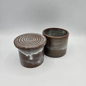 May include: Two-piece ceramic butter dish with a lid and a separate cylindrical container. The dish is dark brown with a white and light blue glaze around the base and on the lid. The lid has a spiral design. The second container is also dark brown with a white and light blue glaze.