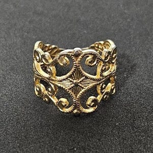 May include: A gold-colored ring with an intricate openwork design. The ring features a repeating pattern of swirling shapes and a central starburst design. The ring is set against a dark background.