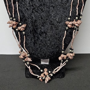 May include: A multi-strand necklace featuring copper-colored shell-shaped beads interspersed with small black and metallic silver beads. The design consists of three strands that drape down, connected by clusters of the shell beads at intervals.